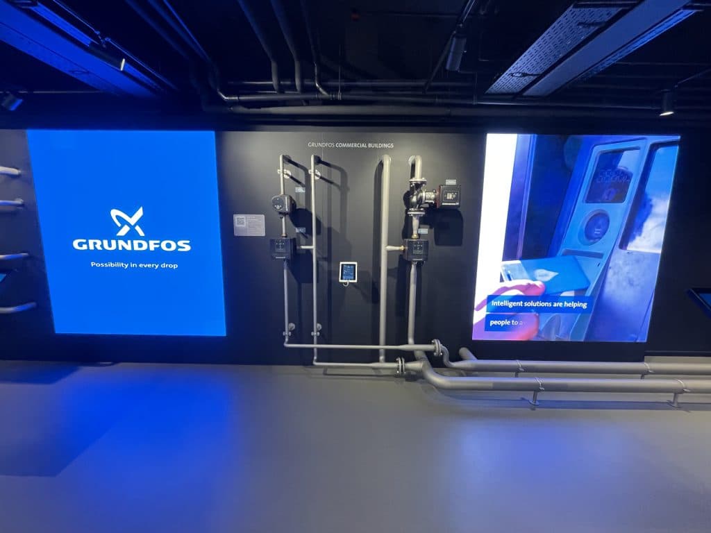 Grundfos Commercial Buildings-installationen med pumper, rør og styringer på en mørk væg ved siden af en stor Grundfos-skærm